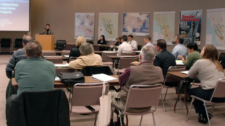 Adults in a classroom setting, all looking forward at a woman presenting a slideshow at the front of the room.