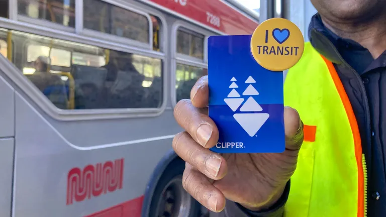 Closeup of a man's hand holding a Clipper card and an "I Heart Transit" pin, as a Muni bus passes by.