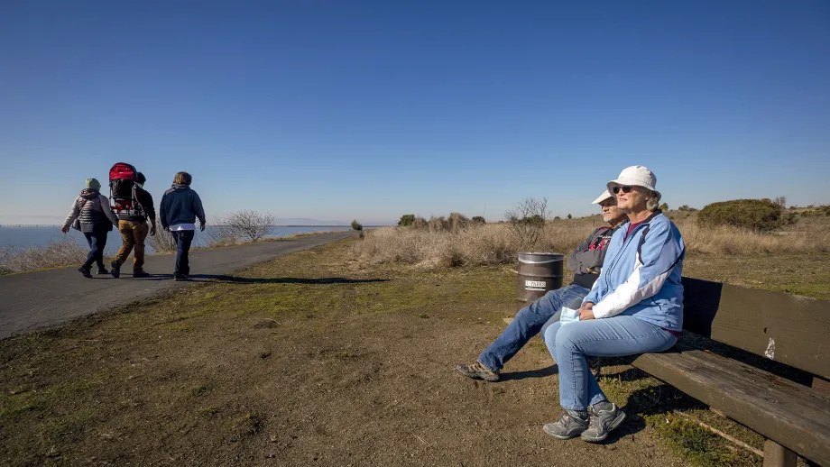 Seniors sitting on a bech as hikers pass by on a path on the Bay Trail.