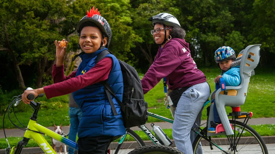A bike-riding family smiles at the camera. A boy wearing a spiked bike helmet is holding a donut and smiling.