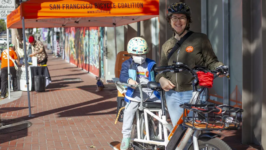 A smiling mom stands with her bicycle with her child in the back seat.