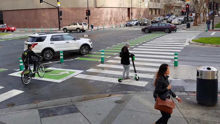 A street intersection with a car, cyclist and scooter in the street, and a pedestrian on the sidewalk.