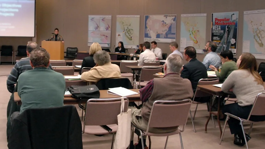 Adults in a classroom setting, all looking forward at a woman presenting a slideshow at the front of the room.