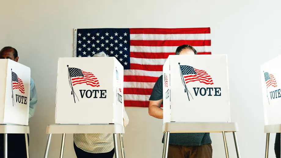 People at voting booths, with an American flag behind them.